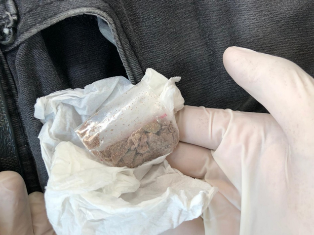Gloved hand holding small bag of brown substance with paper next to gray jeans.